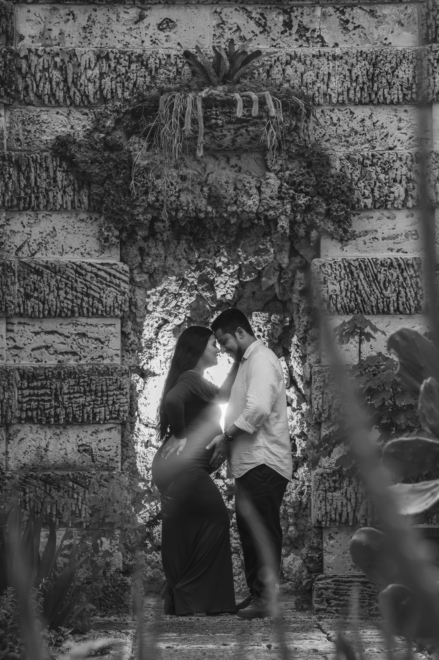 Black and white romantic couple embracing in a stone archway surrounded by lush greenery.