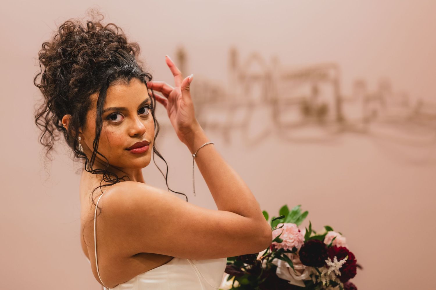 Bride with curly updo holding pink and burgundy bouquet, posing elegantly in a white dress.