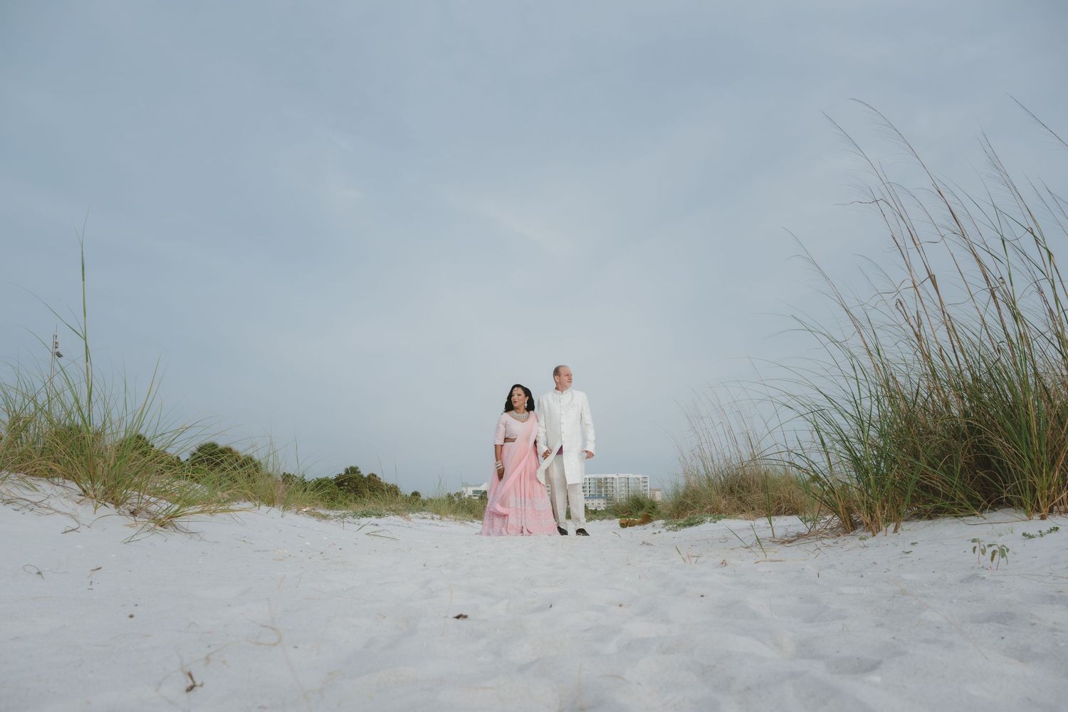 Couple in pink dress and white suit walking on white sand beach dunes with sea grass under cloudy sky.