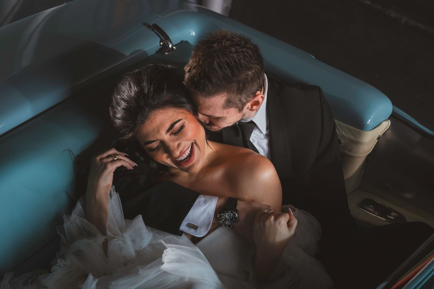 Laughing bride in wedding dress cuddling with groom in tuxedo inside a classic convertible car.