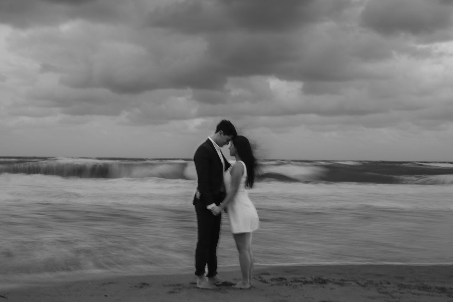 Black and white romantic photo of couple kissing on stormy beach with dramatic crashing ocean waves.