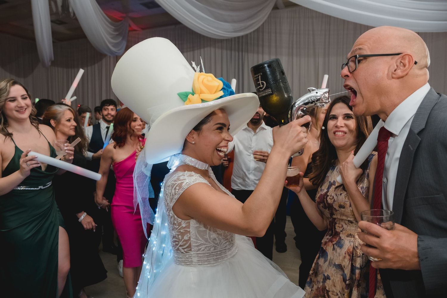 Bride in white lace dress and tall white hat laughing with guests at festive wedding reception party.