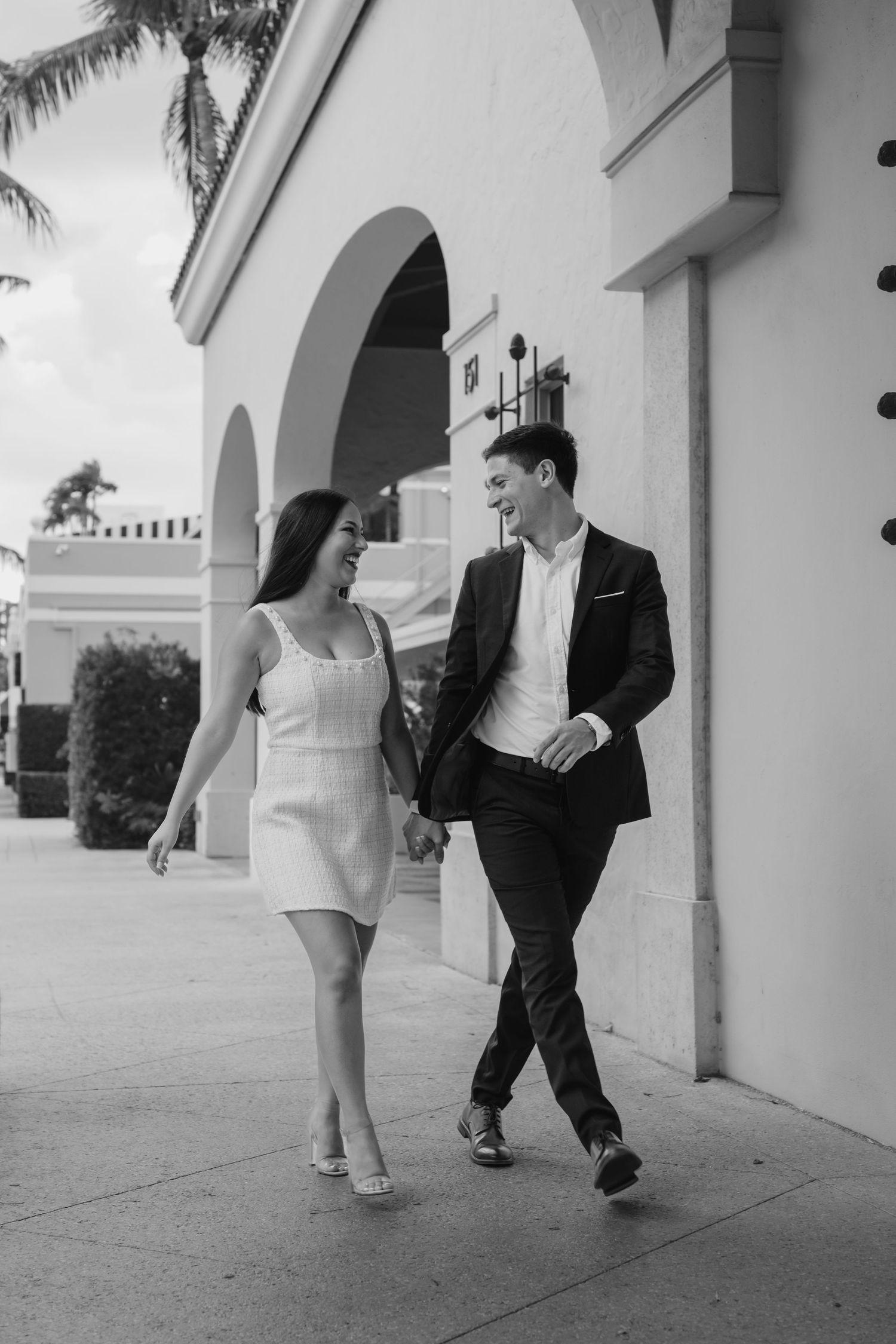 Black and white photo of couple walking hand in hand along arched building exterior.