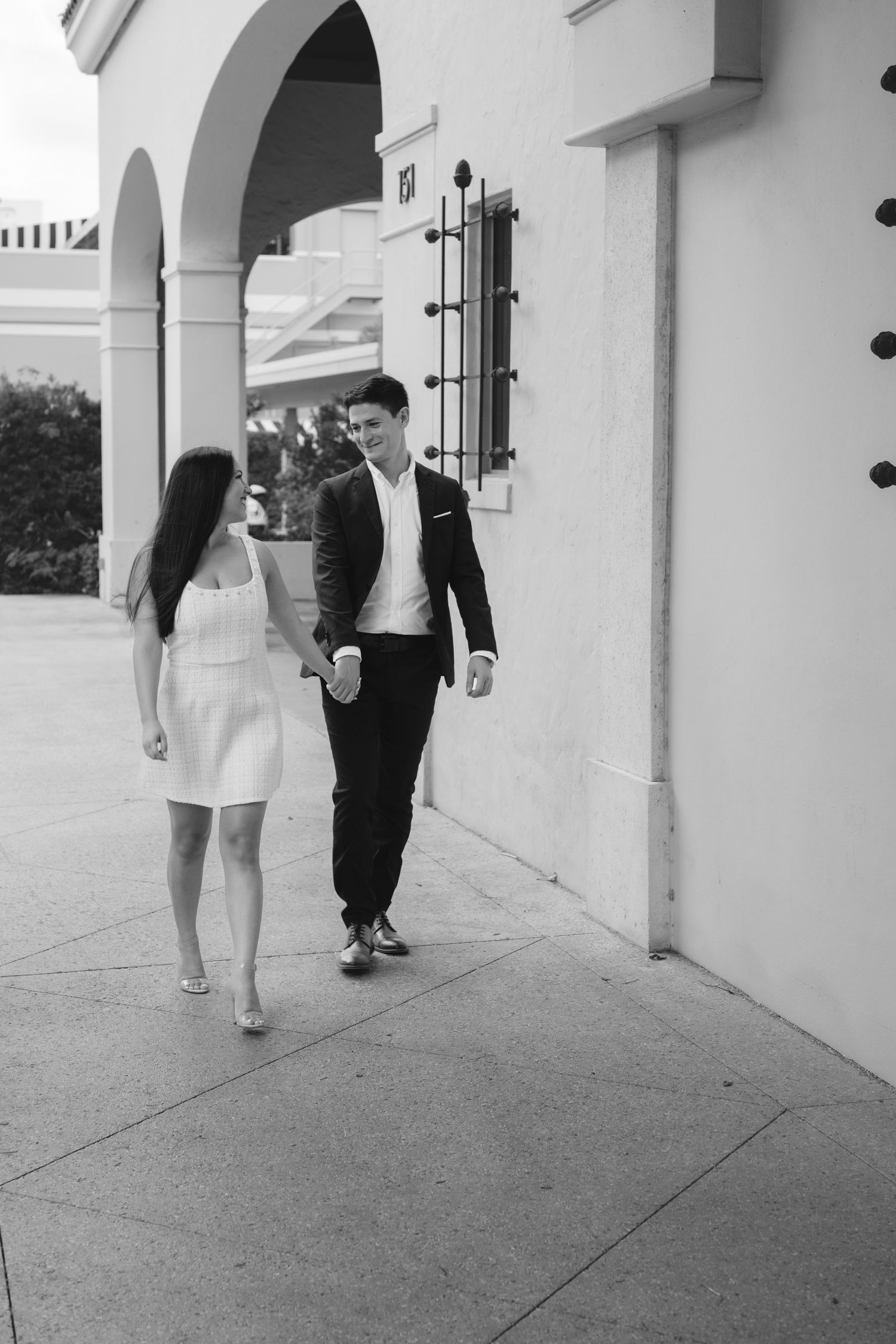 Black and white photo of a couple holding hands and walking along an elegant archway corridor.