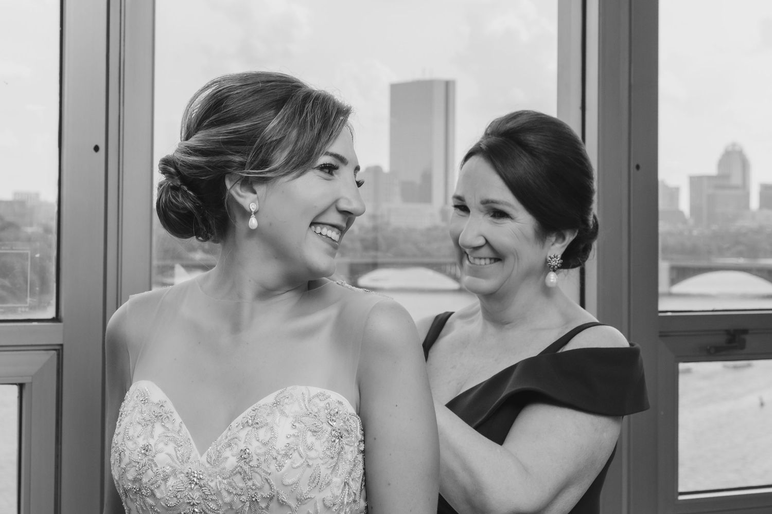 Black and white photo of smiling bride in lace gown with woman by window overlooking city.