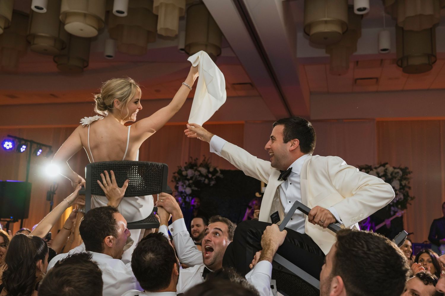 Bride lifted by crowd holding white cloth during lively Jewish wedding hora dance celebration.