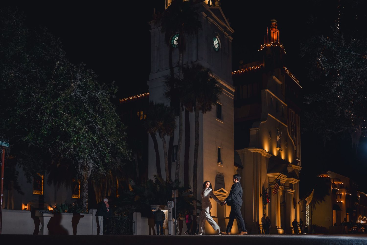 Couple dancing at night outside an illuminated Spanish-style church with palm trees and warm lights.