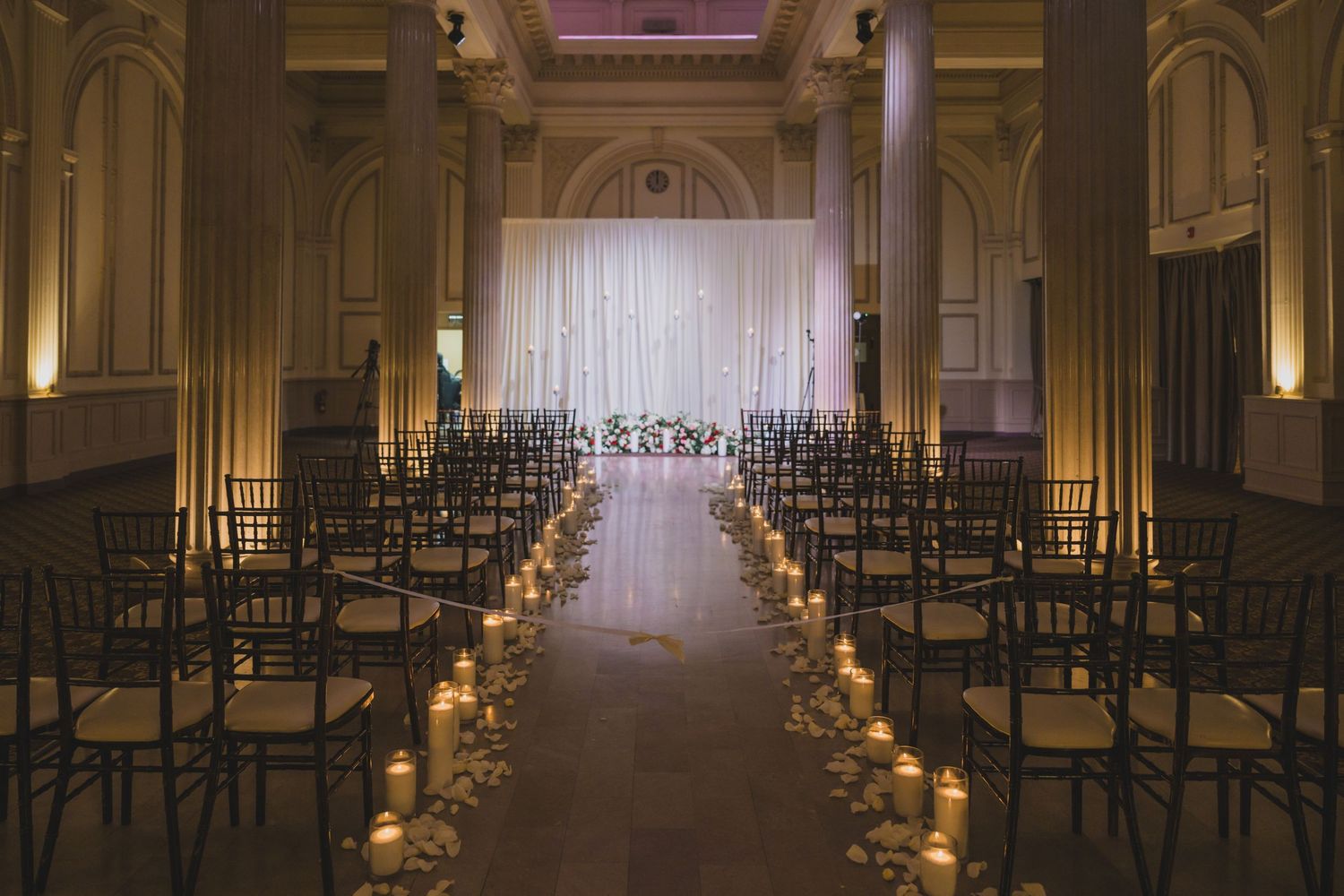 Elegant wedding ceremony aisle with candles, dark chiavari chairs, floral arrangements, and a white draped altar backdrop.