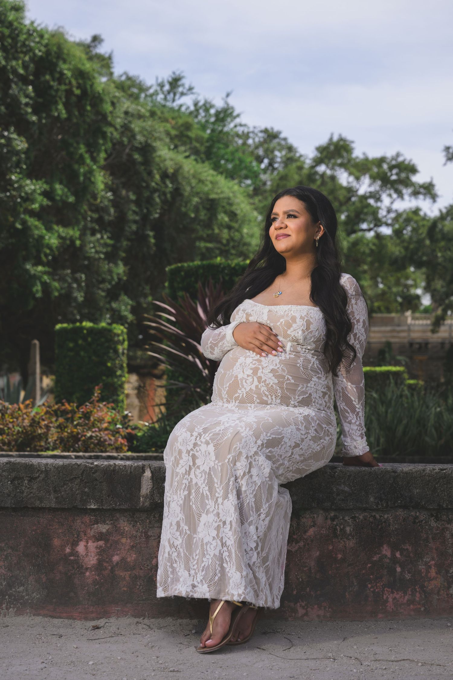 Pregnant woman in white lace gown sitting outdoors, cradling baby bump, surrounded by lush greenery.