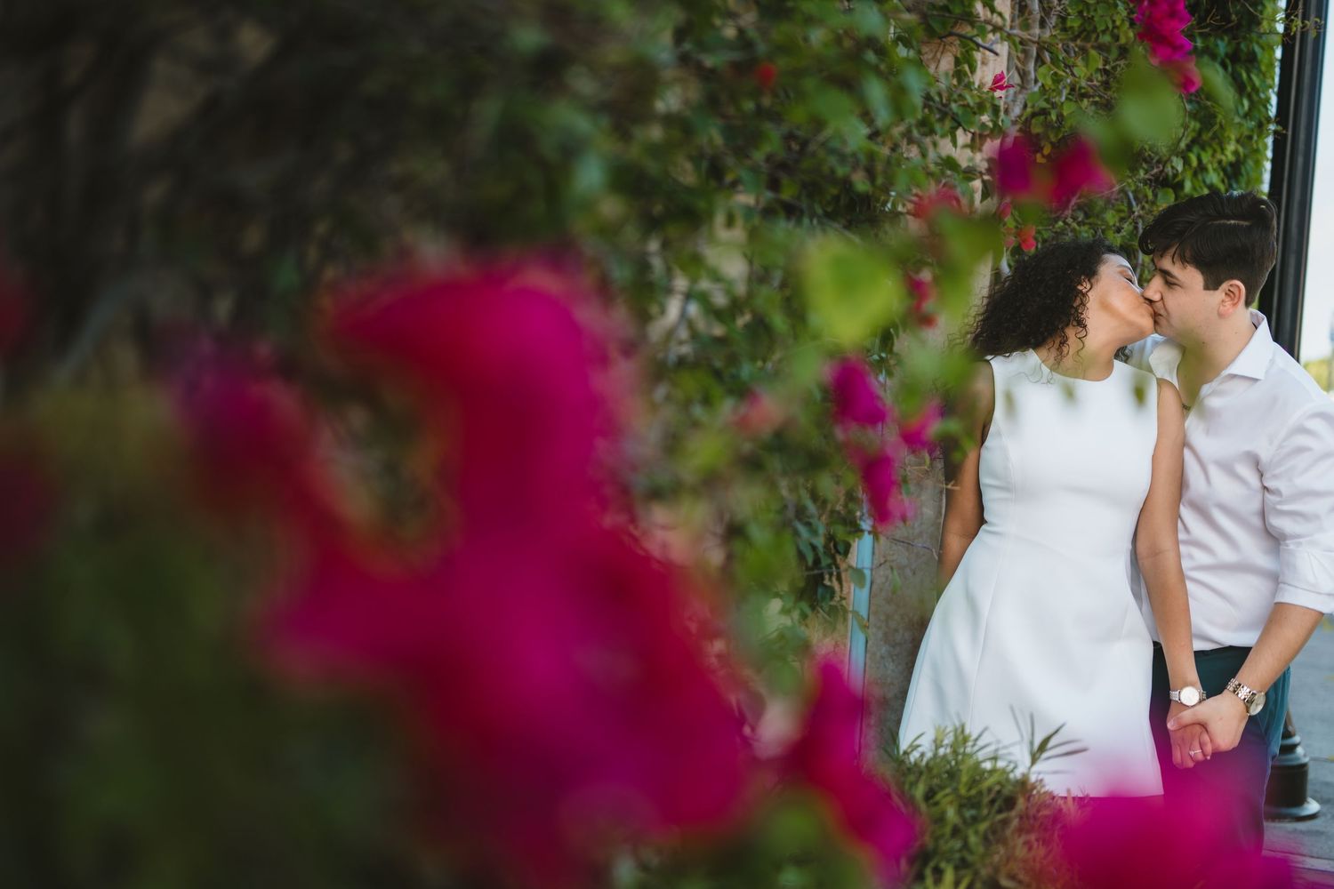 Couple sharing a romantic moment framed by vibrant pink bougainvillea flowers in a garden setting.