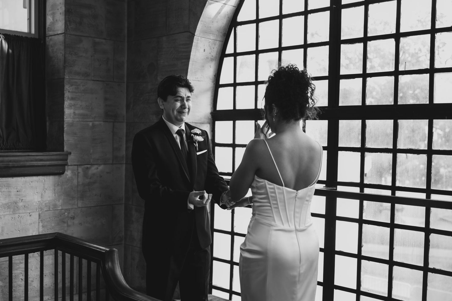 Black and white first look moment between smiling groom and bride near ornate grid window indoors.