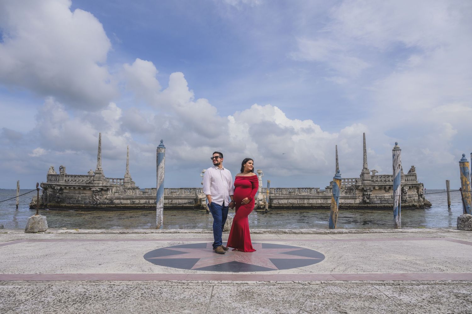 Couple poses on waterfront compass rose with historic Vizcaya Museum statues and cloudy sky behind them.