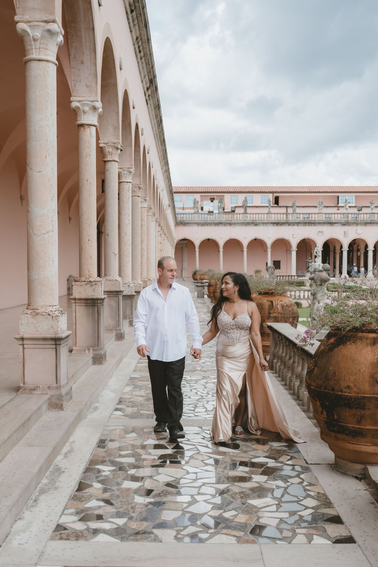 Couple walking hand-in-hand through elegant arched colonnade, woman in gold gown, man in white shirt, at historic venue.