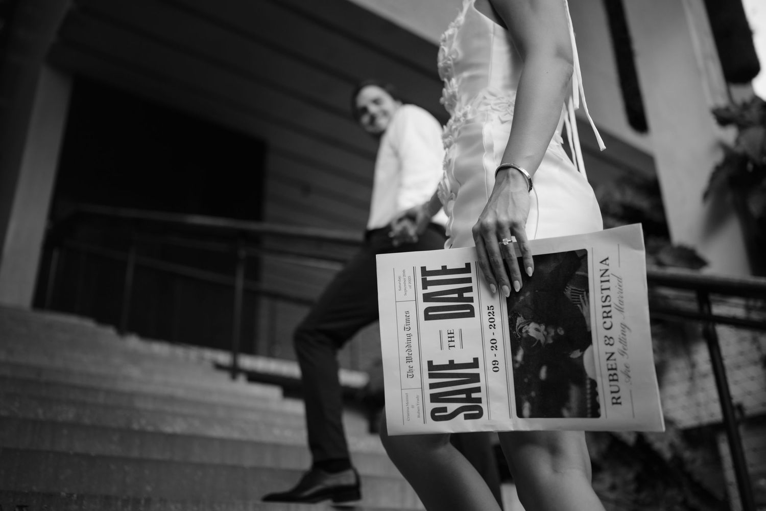 Black and white photo of bride holding a newspaper-style Save the Date card on wedding steps.