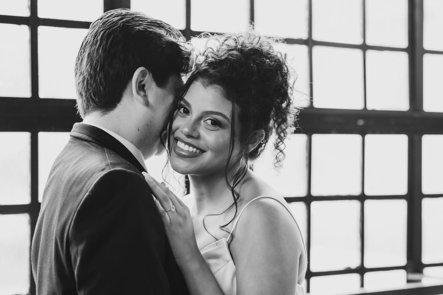 Smiling bride leans close to groom in a romantic black and white wedding portrait by large windows.
