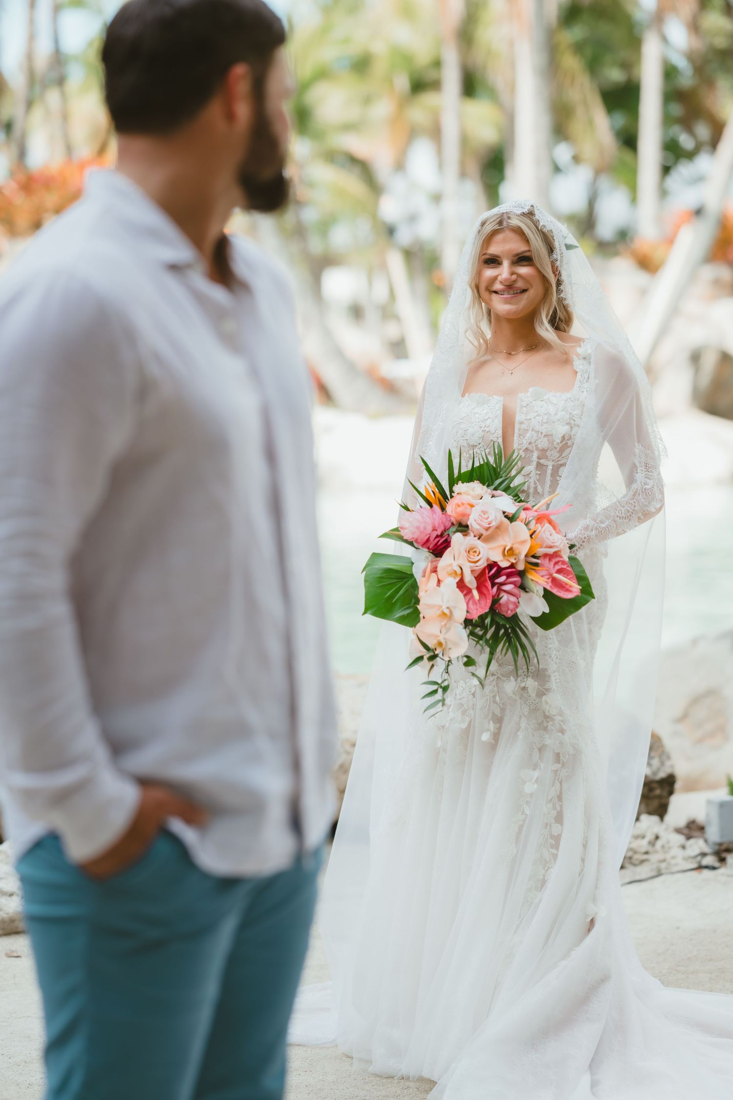 Smiling blonde bride in lace gown holds tropical pink bouquet during outdoor first look.