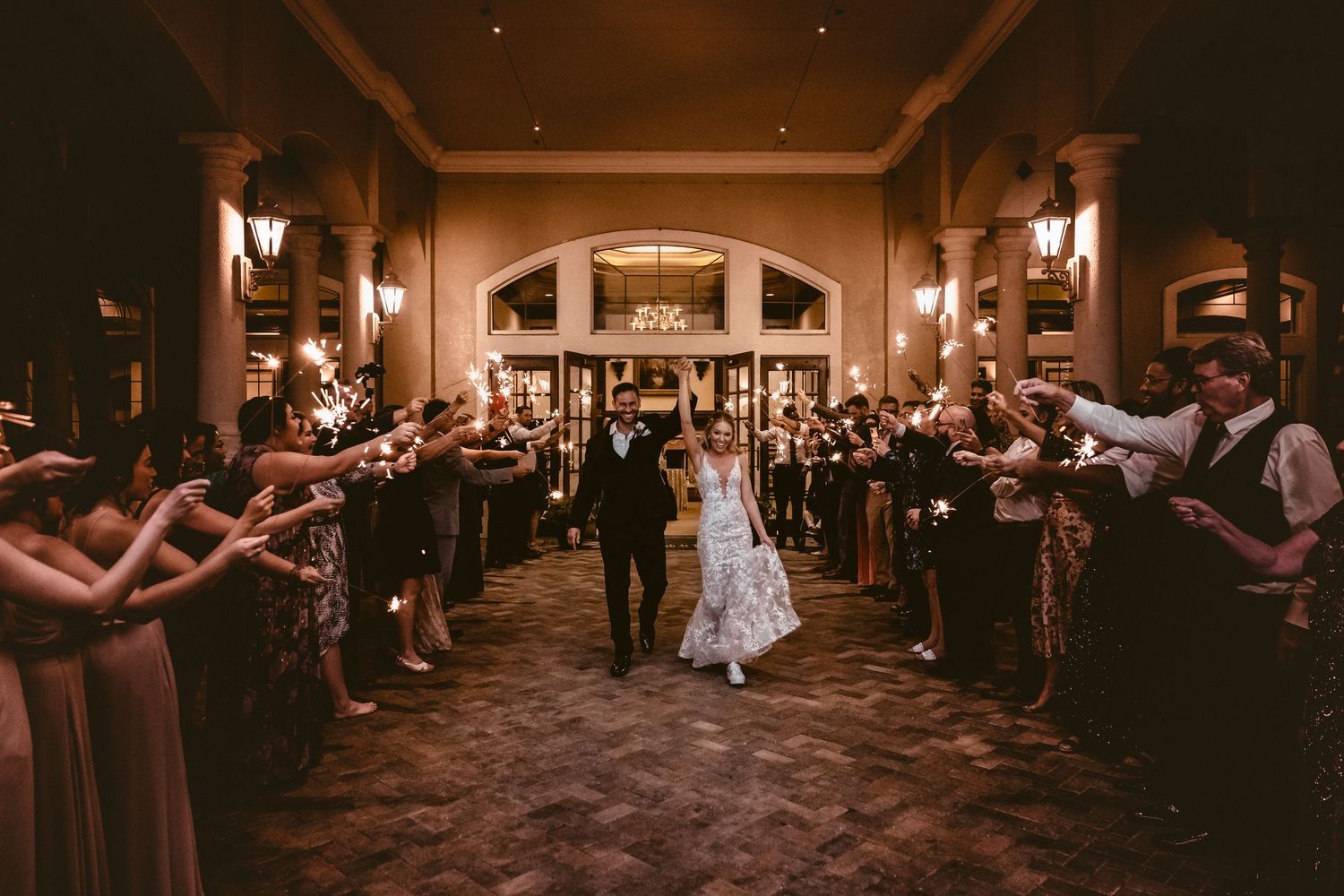 Newlyweds exit wedding venue through sparkler tunnel surrounded by cheering guests.