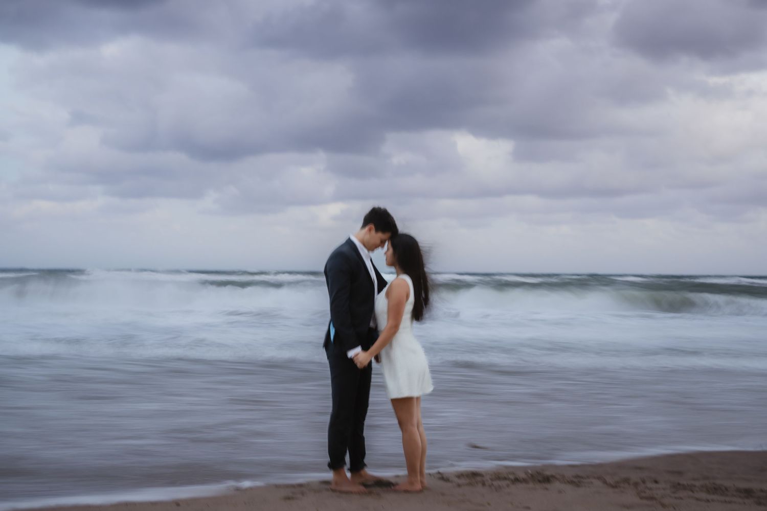 Couple standing barefoot on a beach sharing an intimate moment with dramatic stormy waves.
