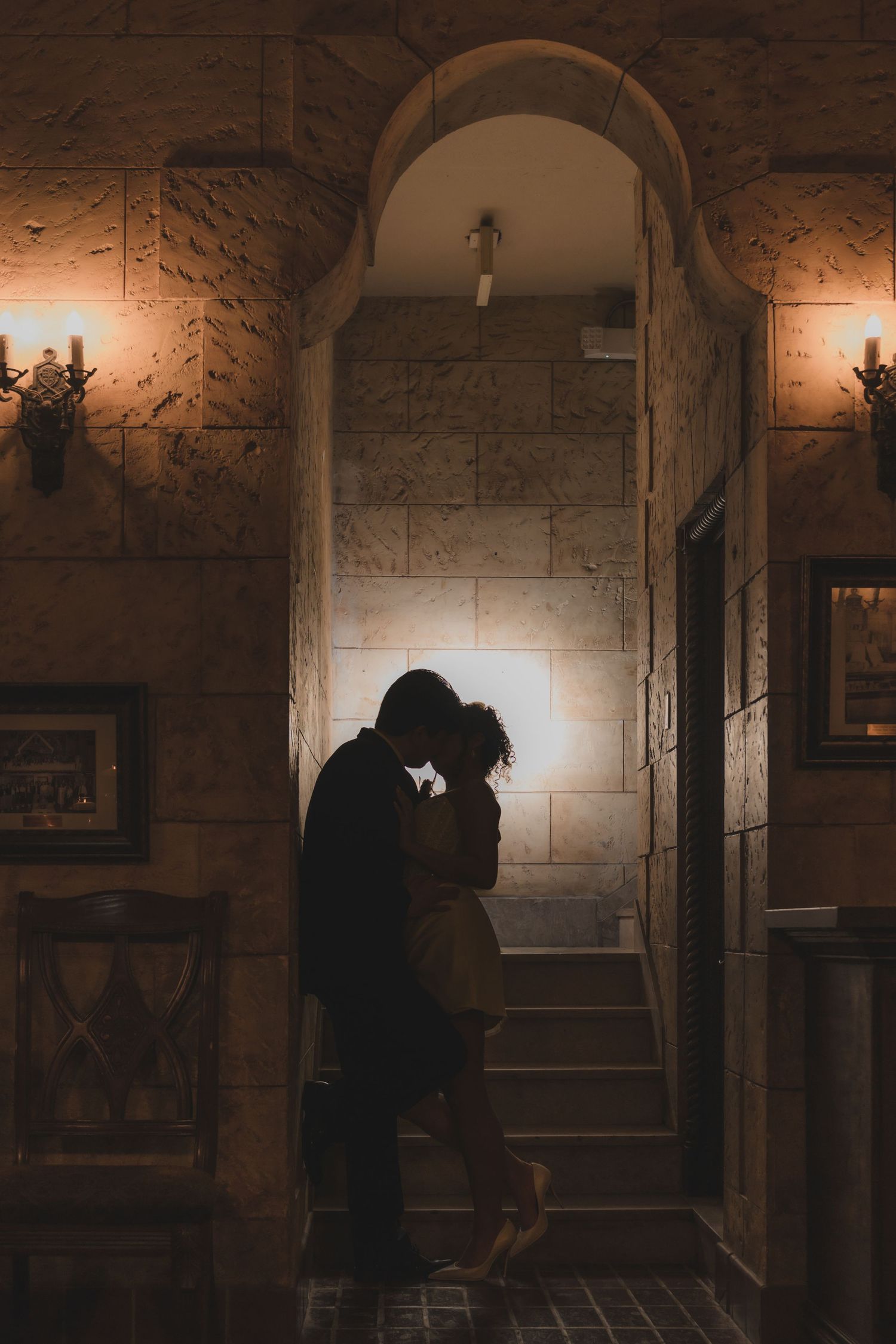 Romantic couple silhouetted in a warmly lit stone archway, sharing an intimate moment in a rustic interior.