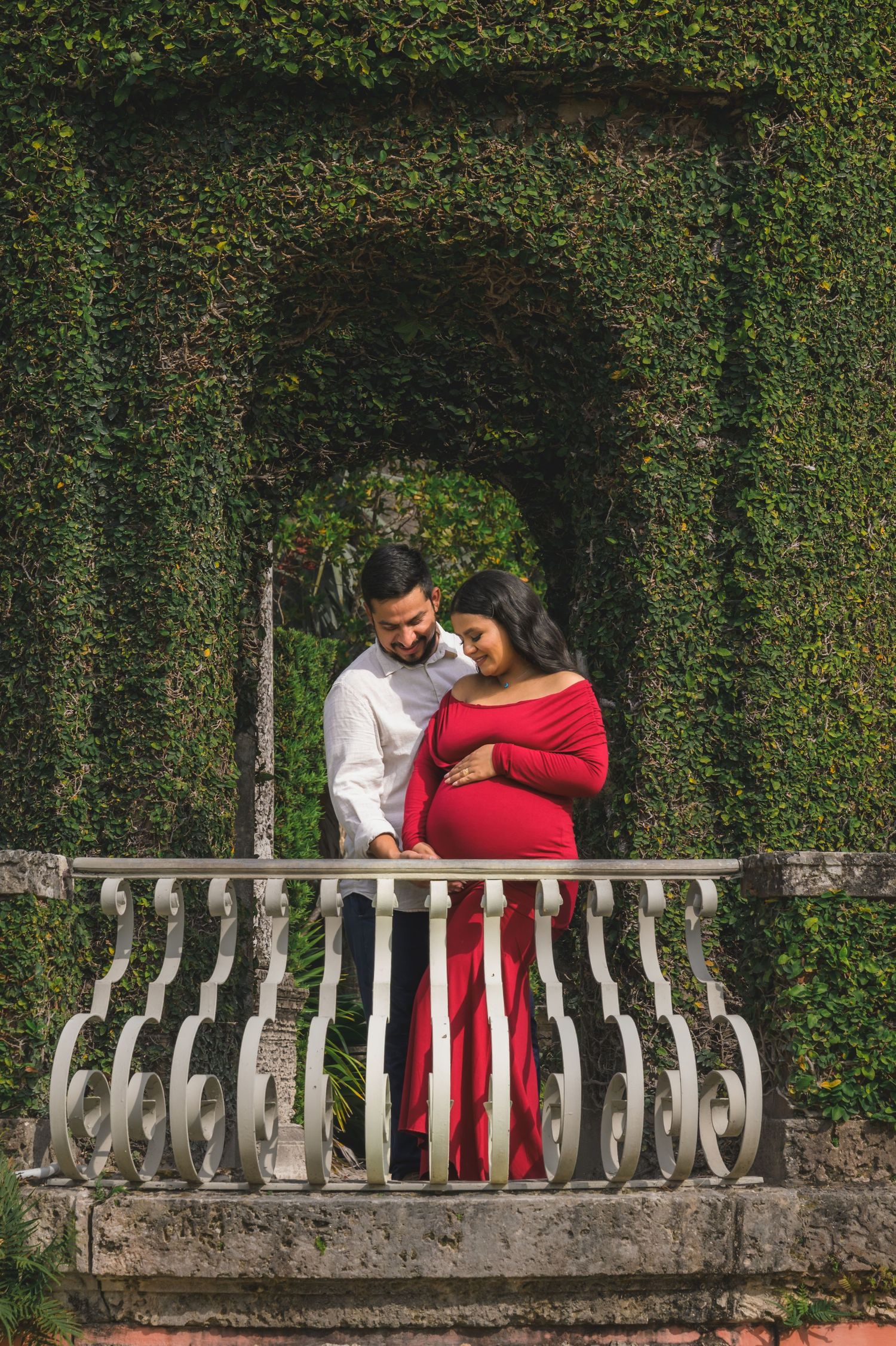 Couple tenderly embracing on balustrade balcony before lush ivy-covered arched hedge wall.