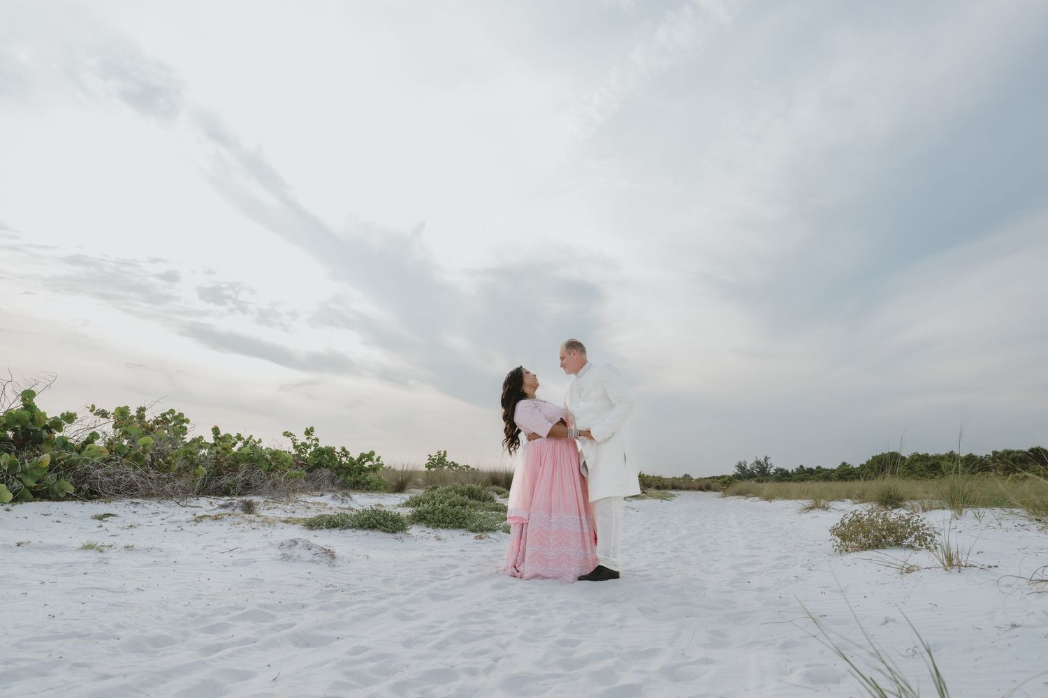 Couple dancing on white sand beach, woman in pink gown and man in white shirt, cloudy sky backdrop.