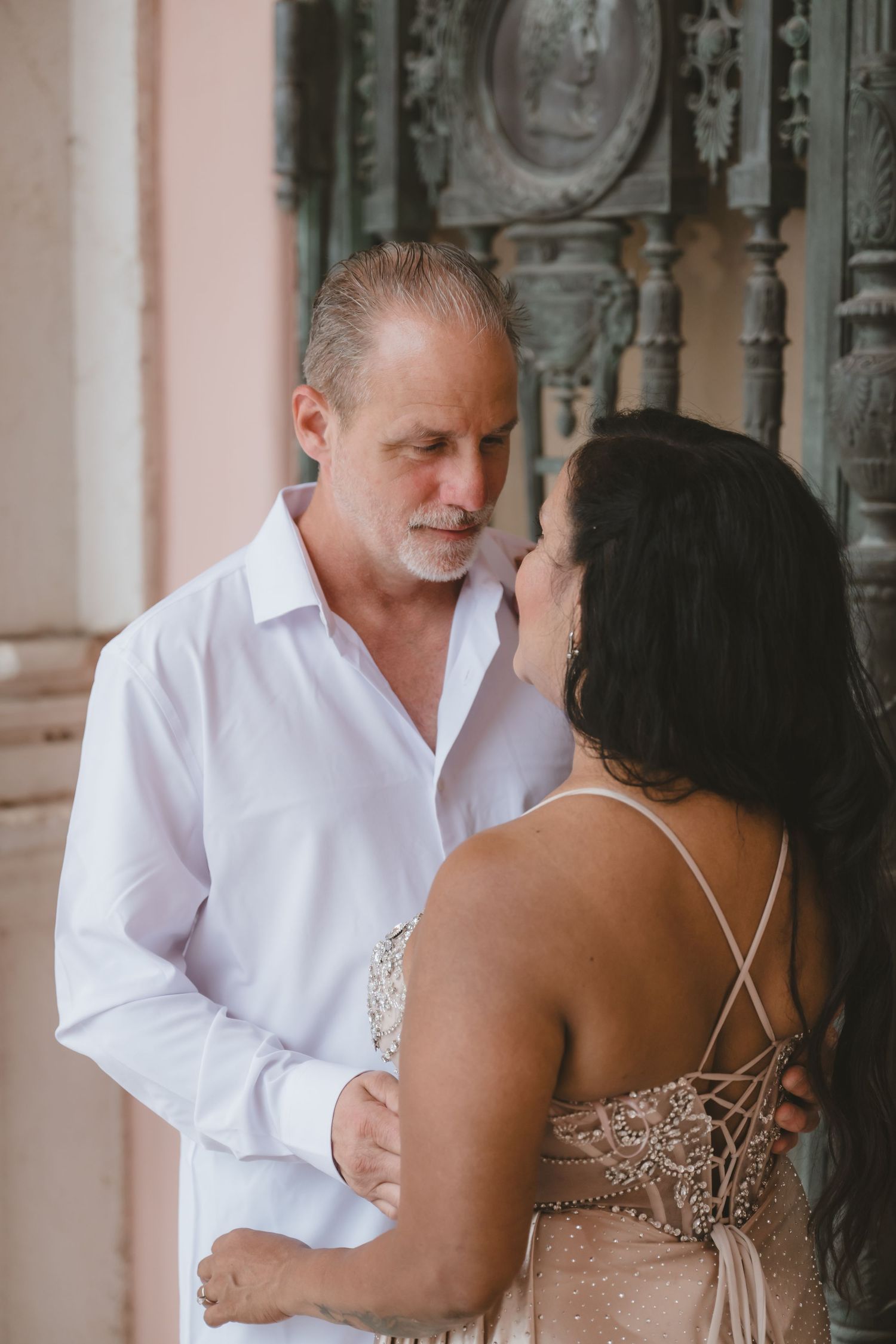 Older couple in romantic embrace, man in white shirt gazing at dark-haired woman in elegant backless beige gown outdoors.