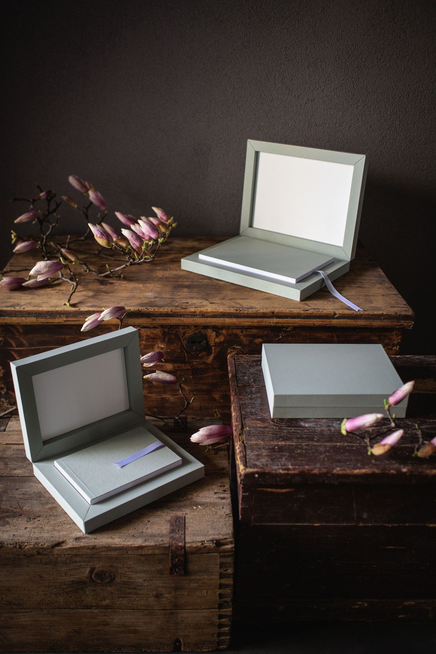 Elegant gray photo boxes with ribbon bookmarks displayed on rustic wooden furniture with pink magnolia flowers.