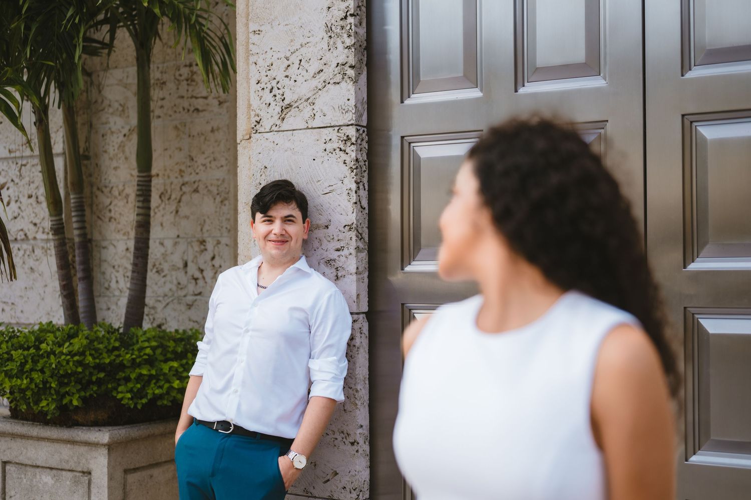 Man in a white shirt smiling at his partner near a large ornate door with tropical foliage.