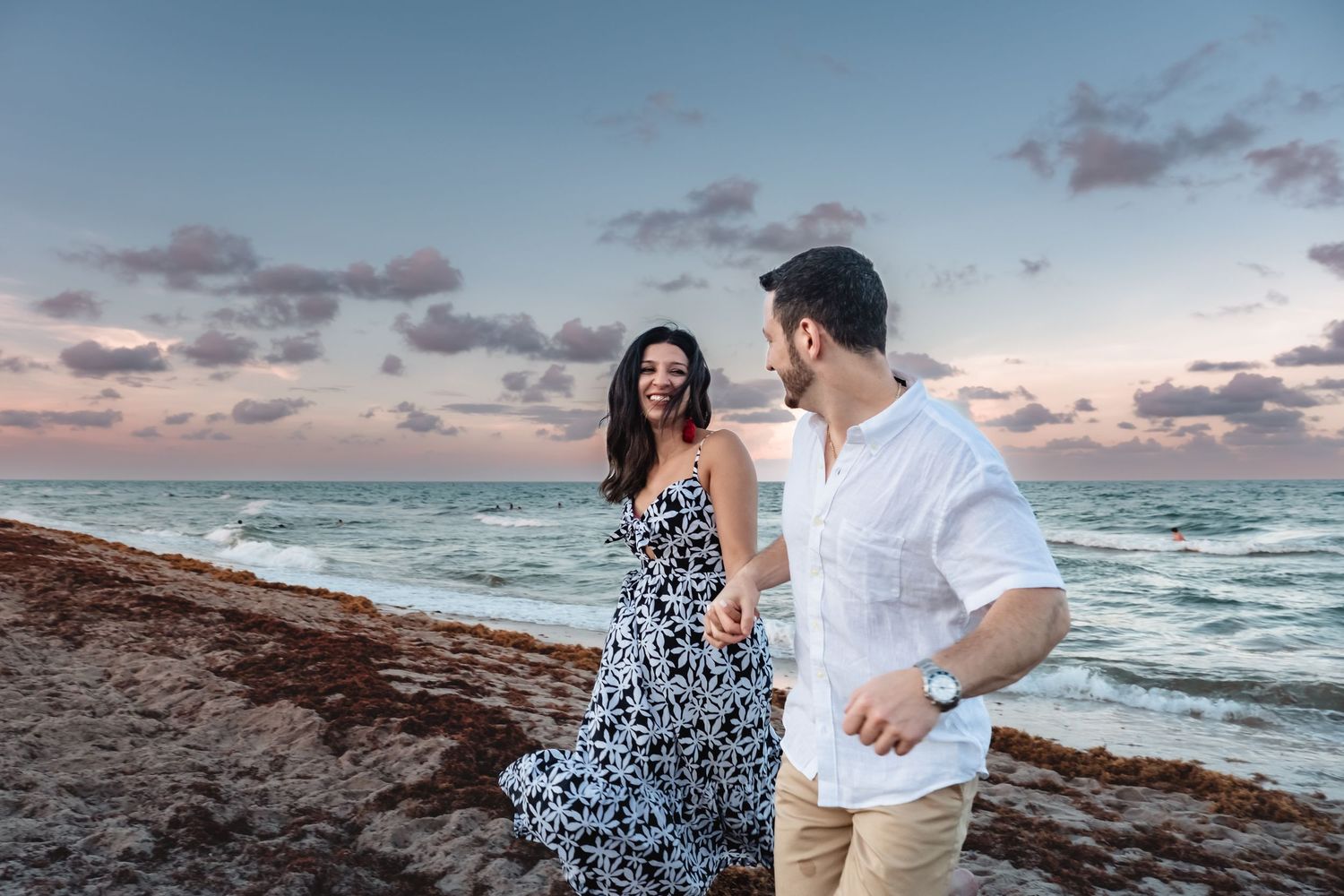 Joyful woman in floral dress holding hands with man while walking on beach at dusk.