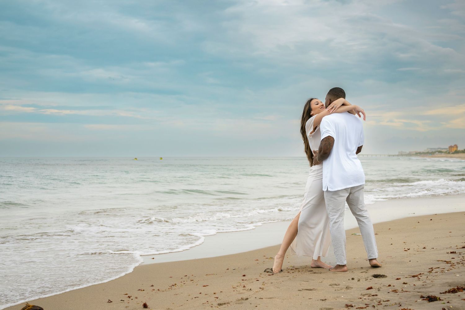 Couple embracing romantically on a sandy beach with ocean waves and cloudy sky backdrop.