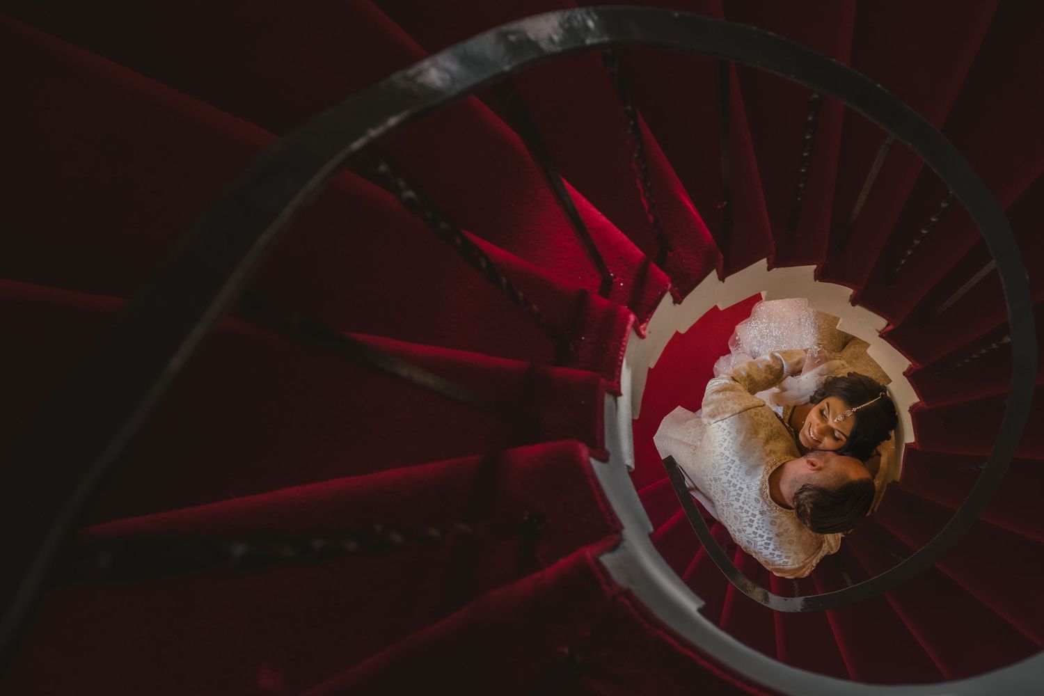 Aerial view of bride and groom embracing at the center of a dramatic red spiral staircase.