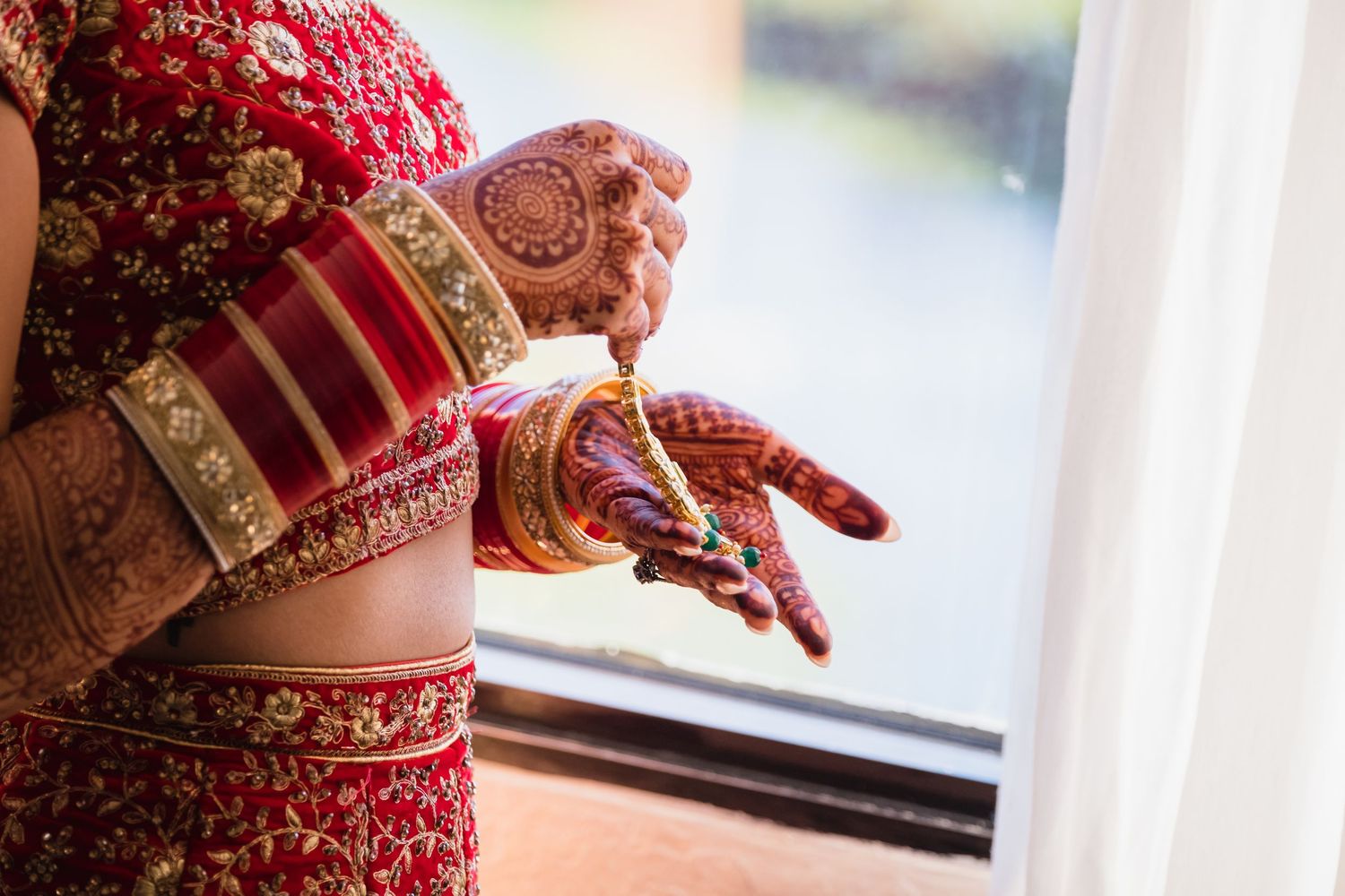 Indian bride in red lehenga adjusting gold bangles and mehndi-adorned hands by window.
