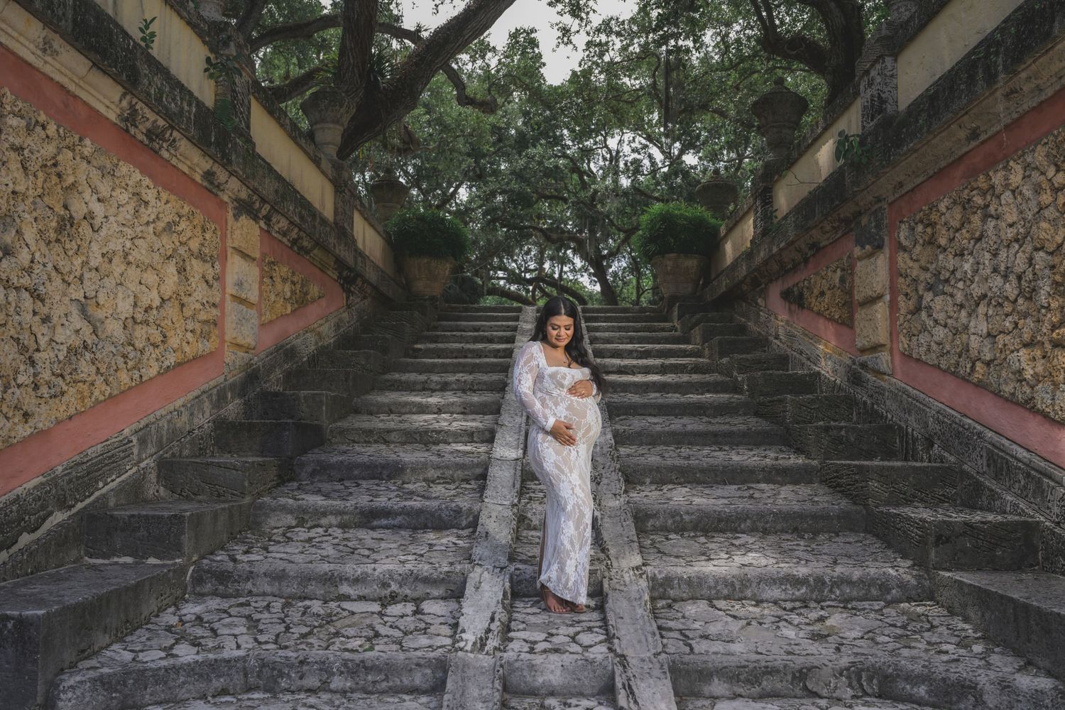 Bride in a white lace gown standing gracefully on stone stairs surrounded by trees and colorful walls.