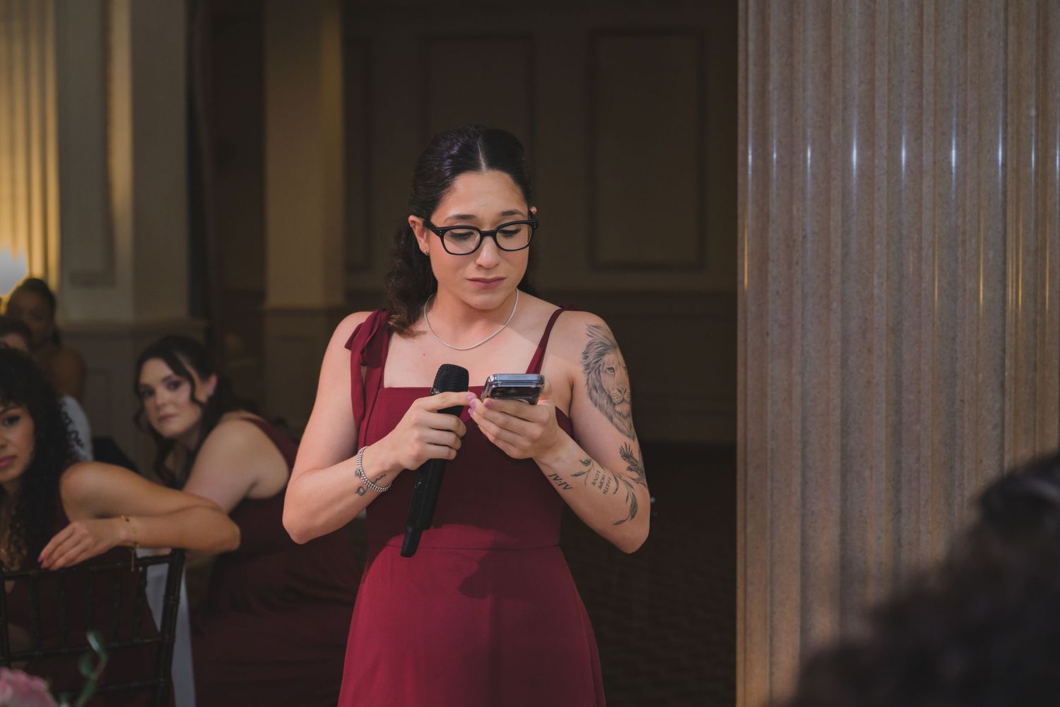 Woman in red dress holding microphone and phone at indoor event, wearing glasses and tattoos.