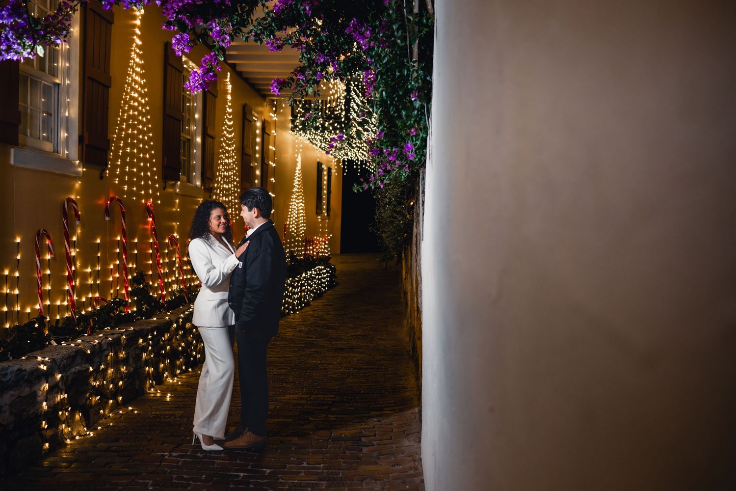 Couple sharing a romantic moment in a charming alley adorned with warm string lights and purple flowers at night.