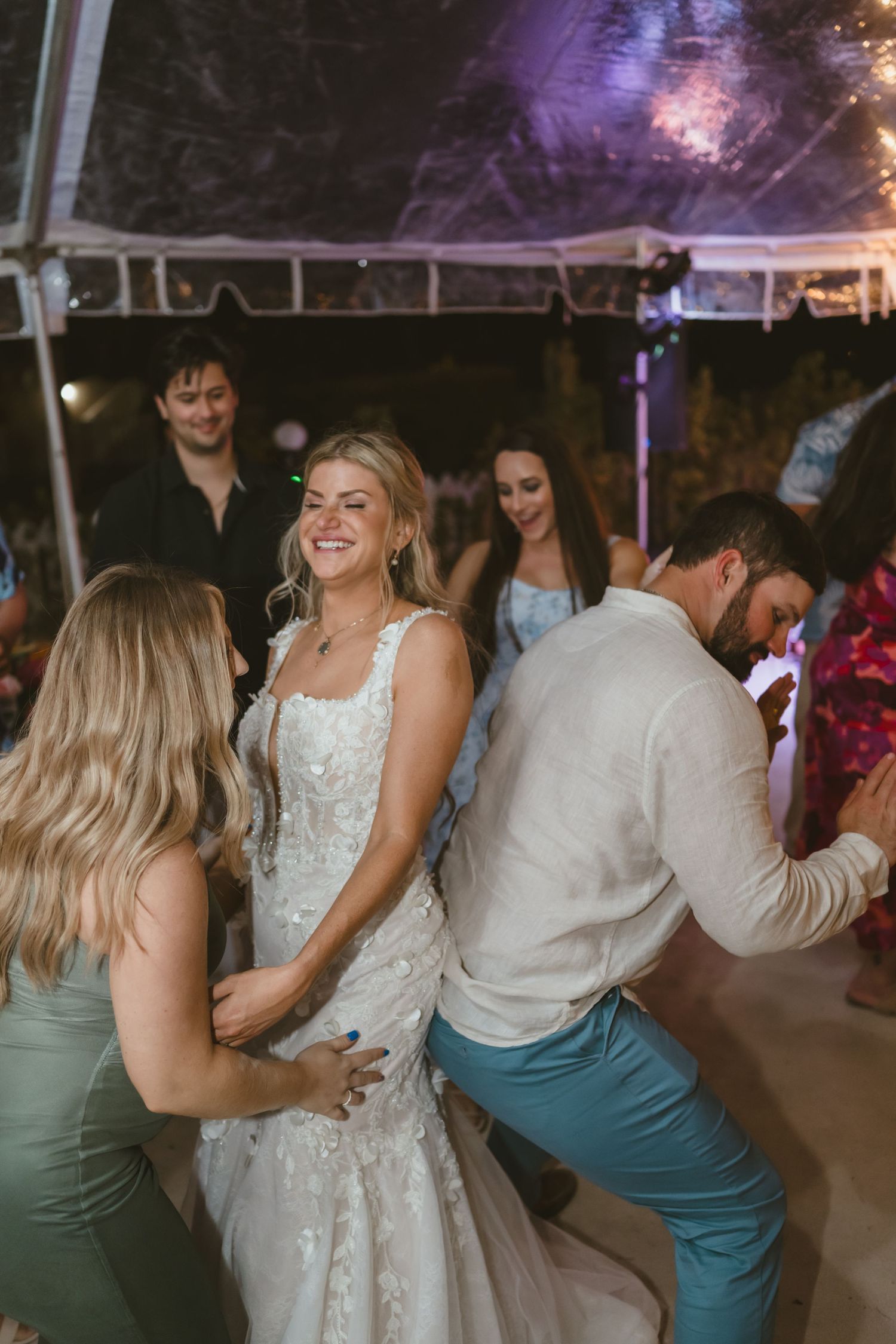 Joyful bride in lace gown dances with guests under a tent at wedding reception.