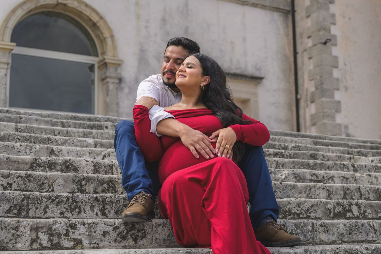 Couple sitting on stone steps, woman in red dress leaning against man in white shirt, romantic outdoor portrait.