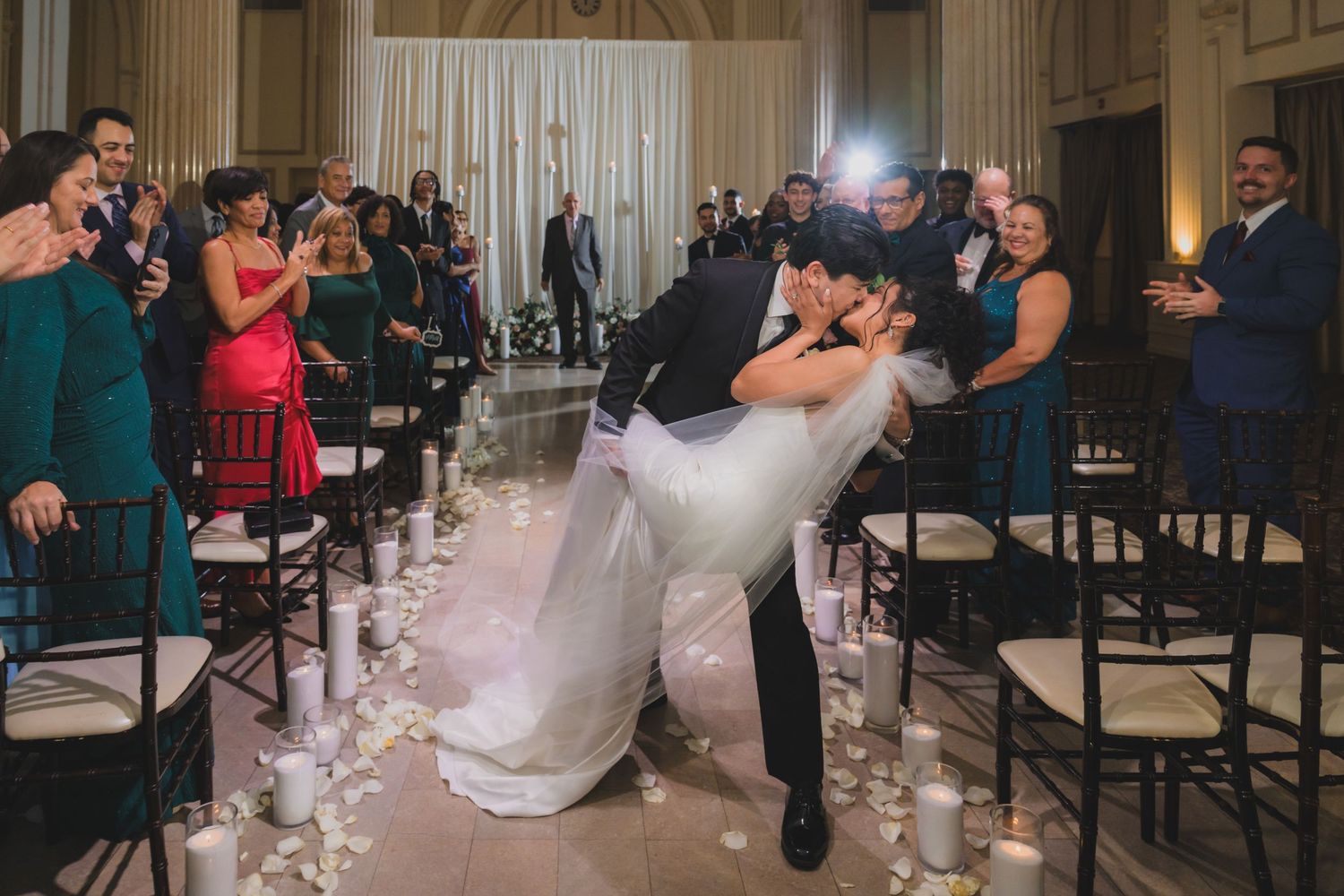 Groom dips bride for a romantic kiss at wedding ceremony aisle lined with candles and rose petals.