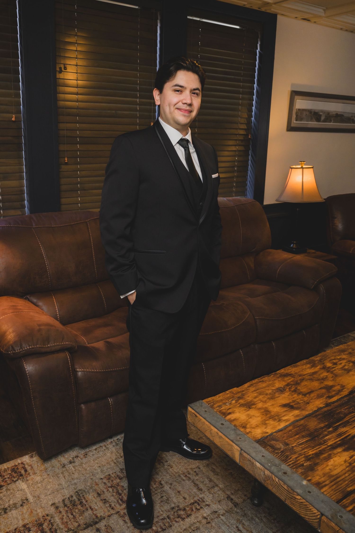Groom in black tuxedo smiling confidently in elegant lounge with leather sofas and warm lighting.