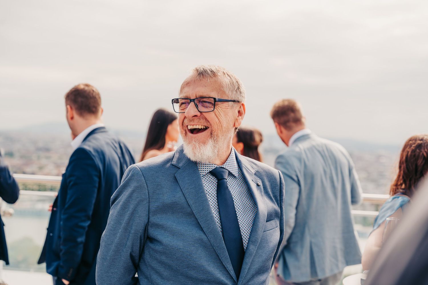 Ein glücklicher älterer Mann mit grauem Bart und Brille lacht bei einem Hochzeits-Empfang im Freien.