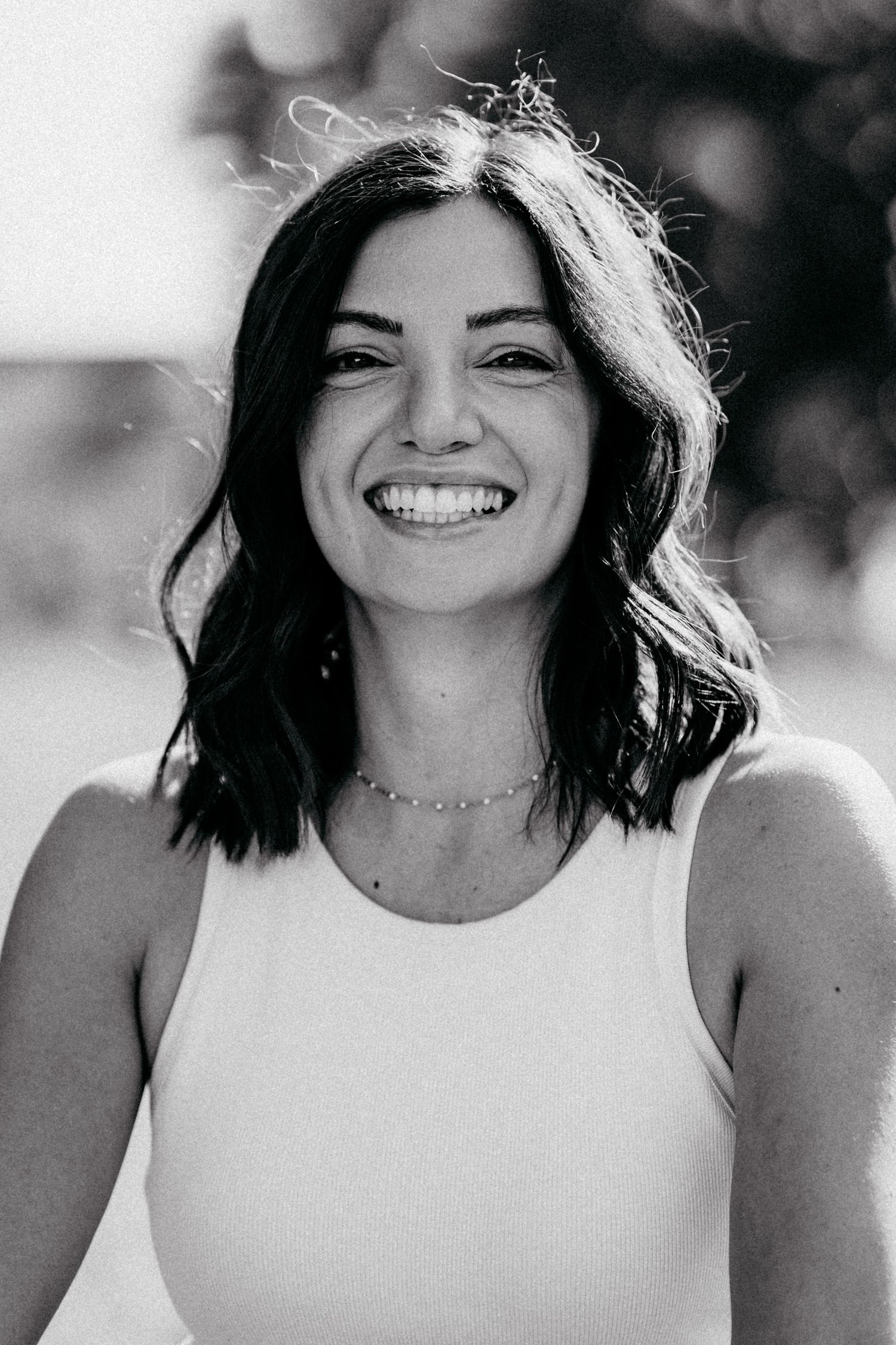 Black and white portrait of a smiling young woman with dark wavy hair wearing a white sleeveless top and delicate necklace.
