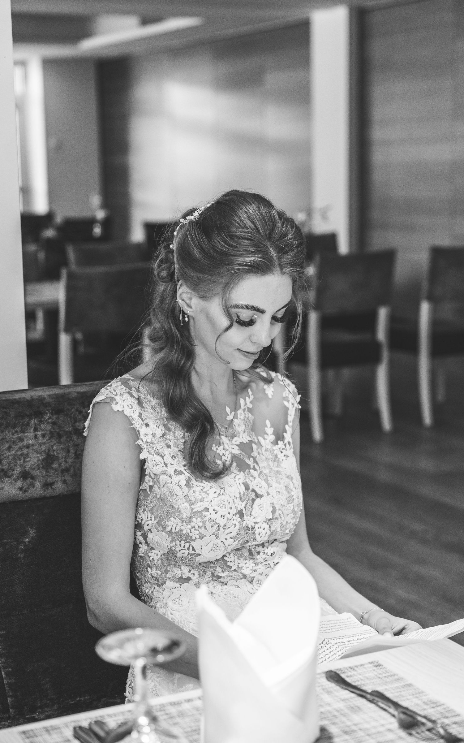 Elegante Braut in einem Spitzenhochzeitskleid sitzt an einem Tisch und liest in einer Schwarz-Weiß-Fotografie.