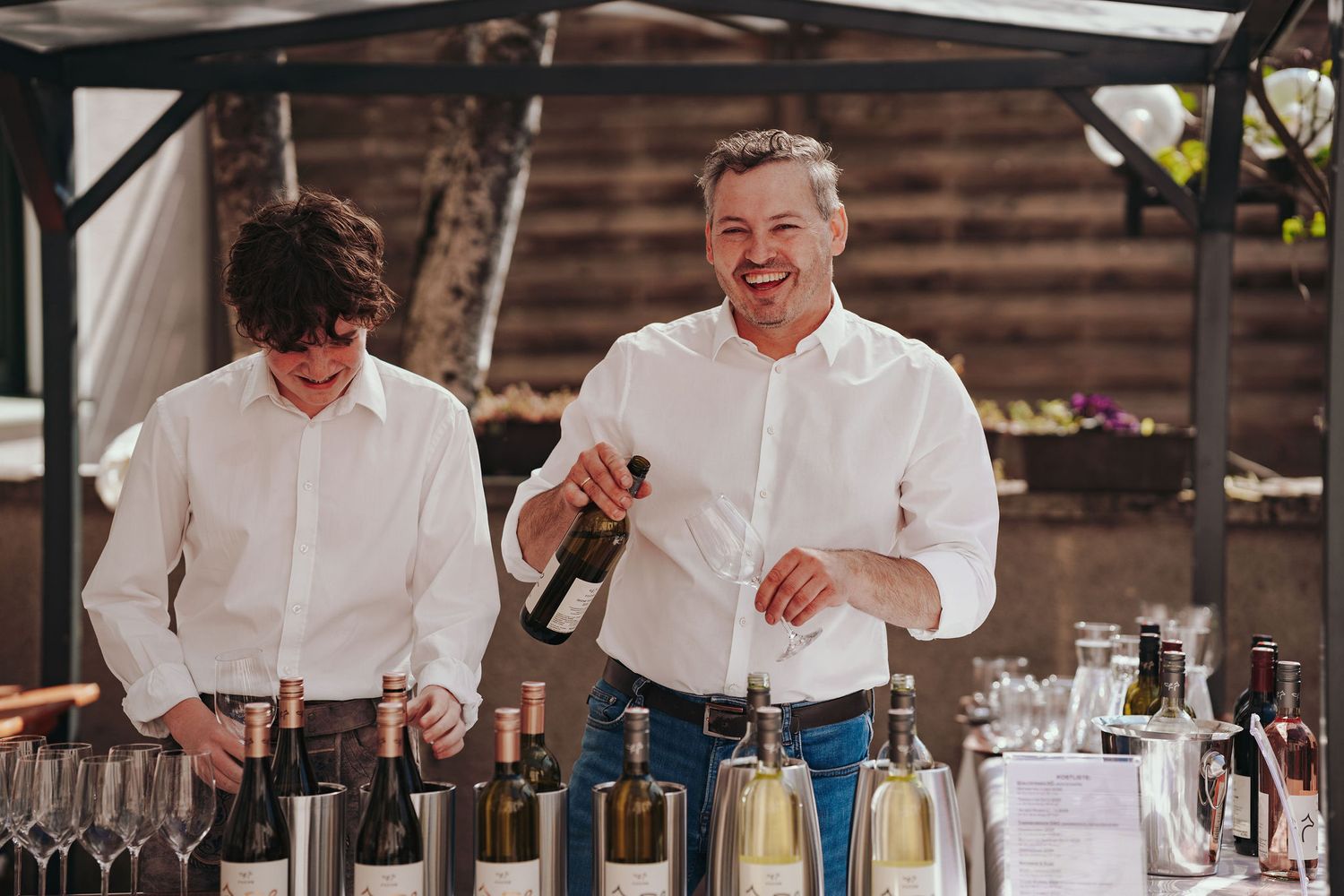 Zwei Winzer in weißen Hemden lachen, während sie bei einer Outdoor-Eventbar Wein ausschenken, mit ausgestellten Flaschen im Hintergrund.