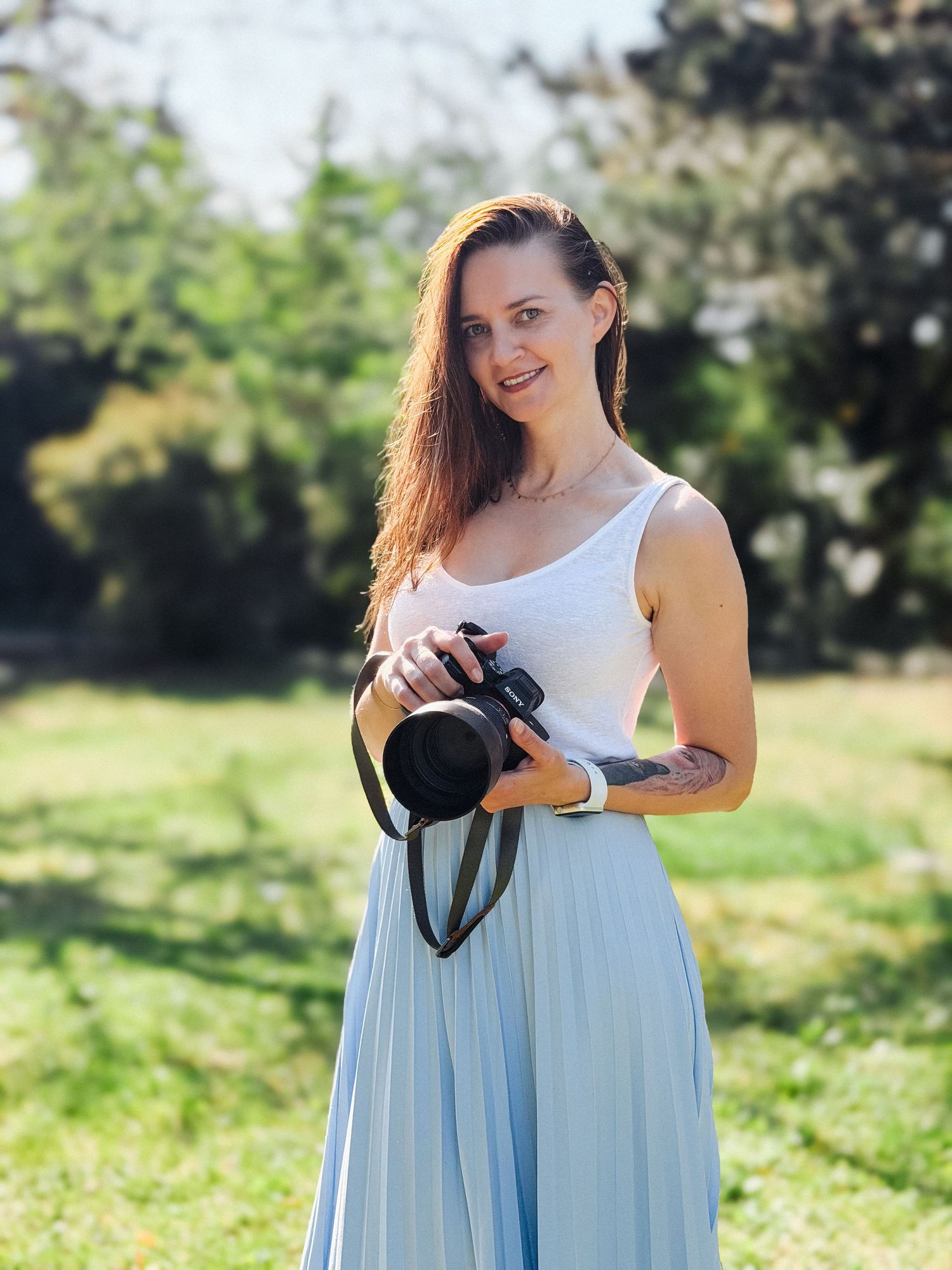 Woman photographer smiling outdoors holding camera, wearing white top and light blue skirt, surrounded by green trees.