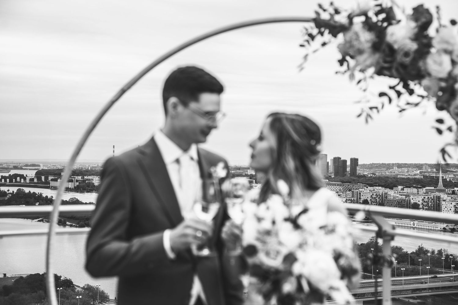 Schwarz-Weiß-Foto einer Braut und eines Bräutigams, die unter einem Blumenbogen über den Dächern Wiens mit Champagner anstoßen.