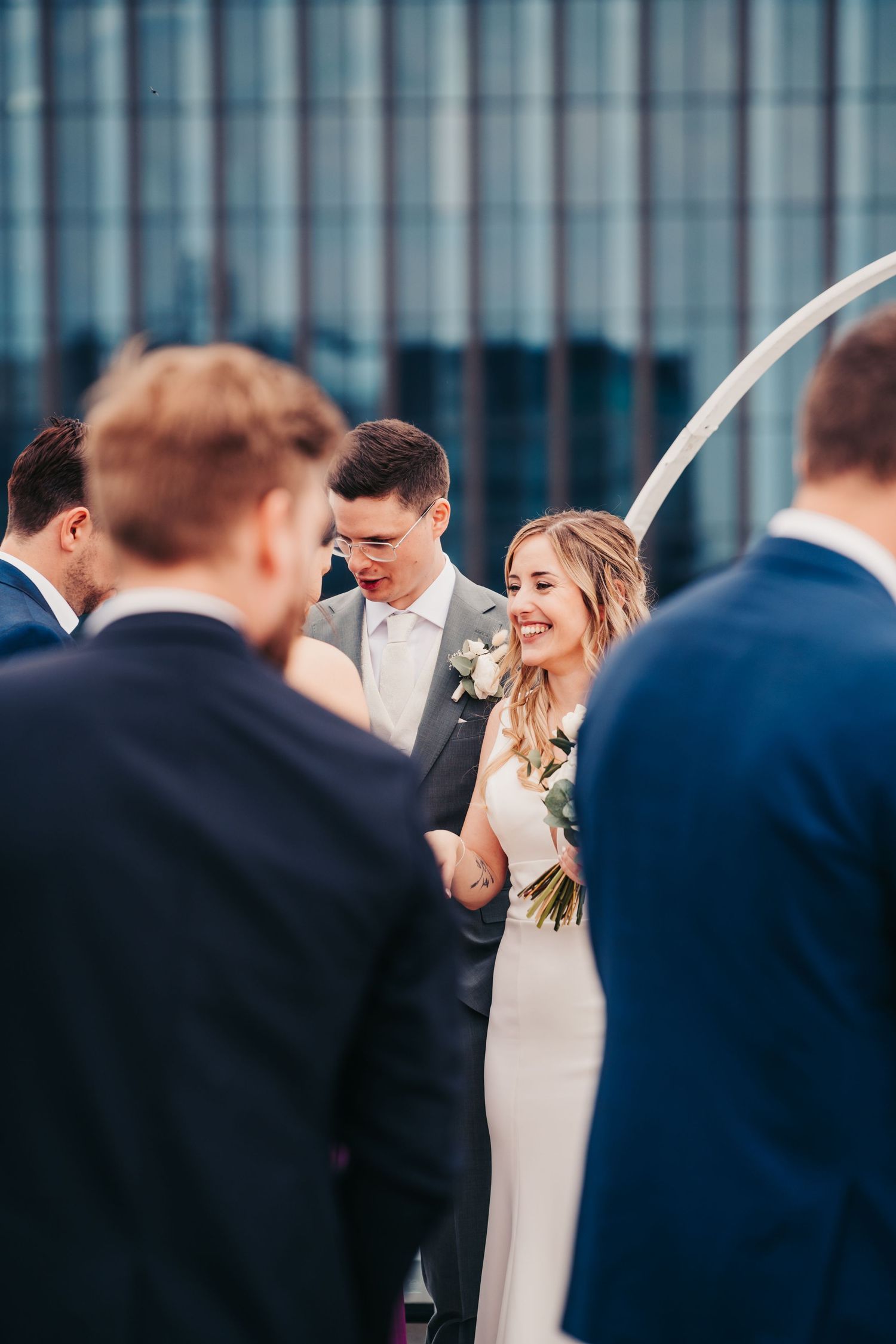 Braut mit Brautstrauß, Bräutigam blickt nach unten, Dachterrassen-Hochzeit mit Gästen im Hintergrund.
