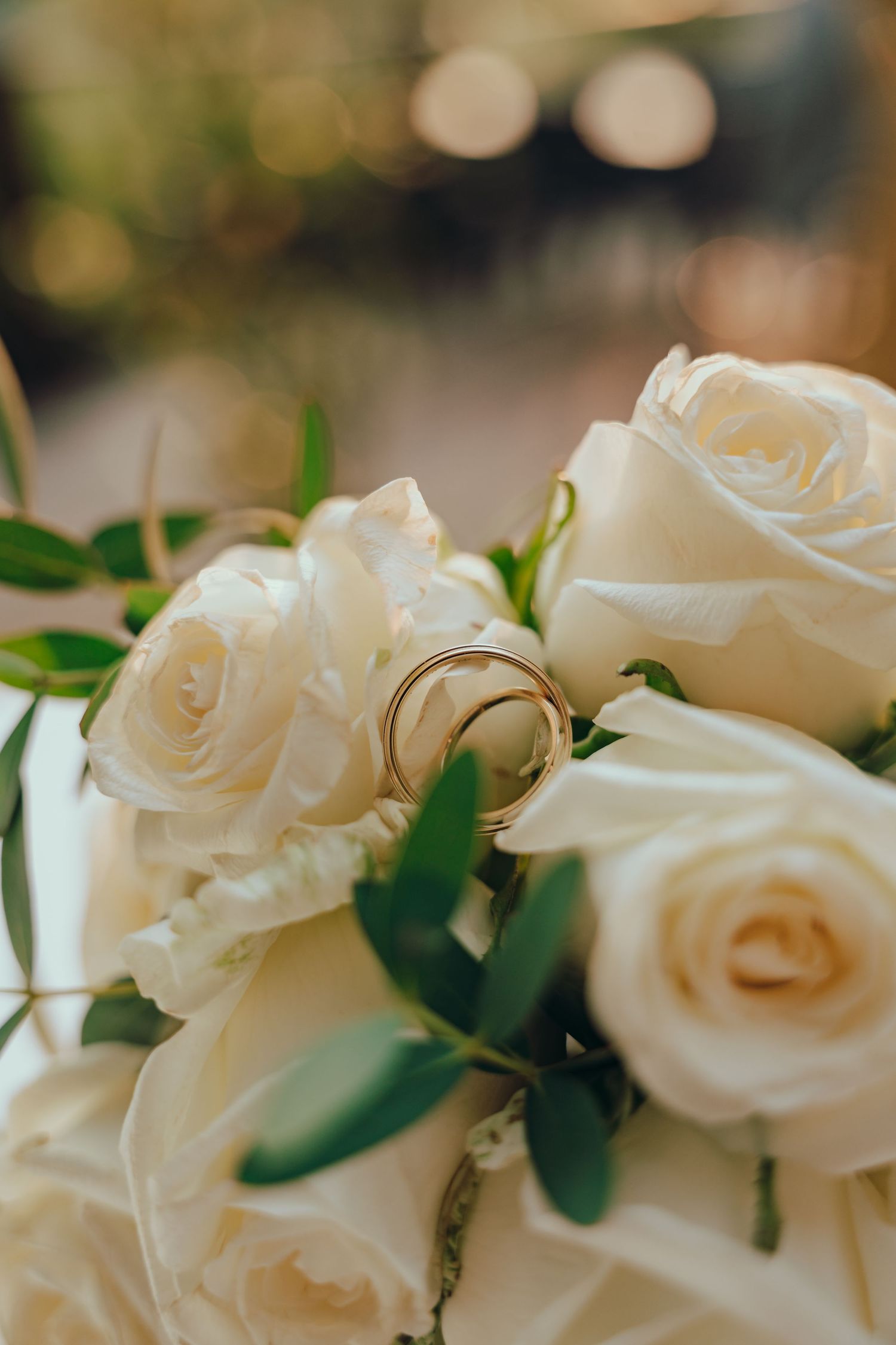 Goldene Eheringe liegen auf einem weißen Rosenbrautstrauß mit grünen Blättern, Nahaufnahme.