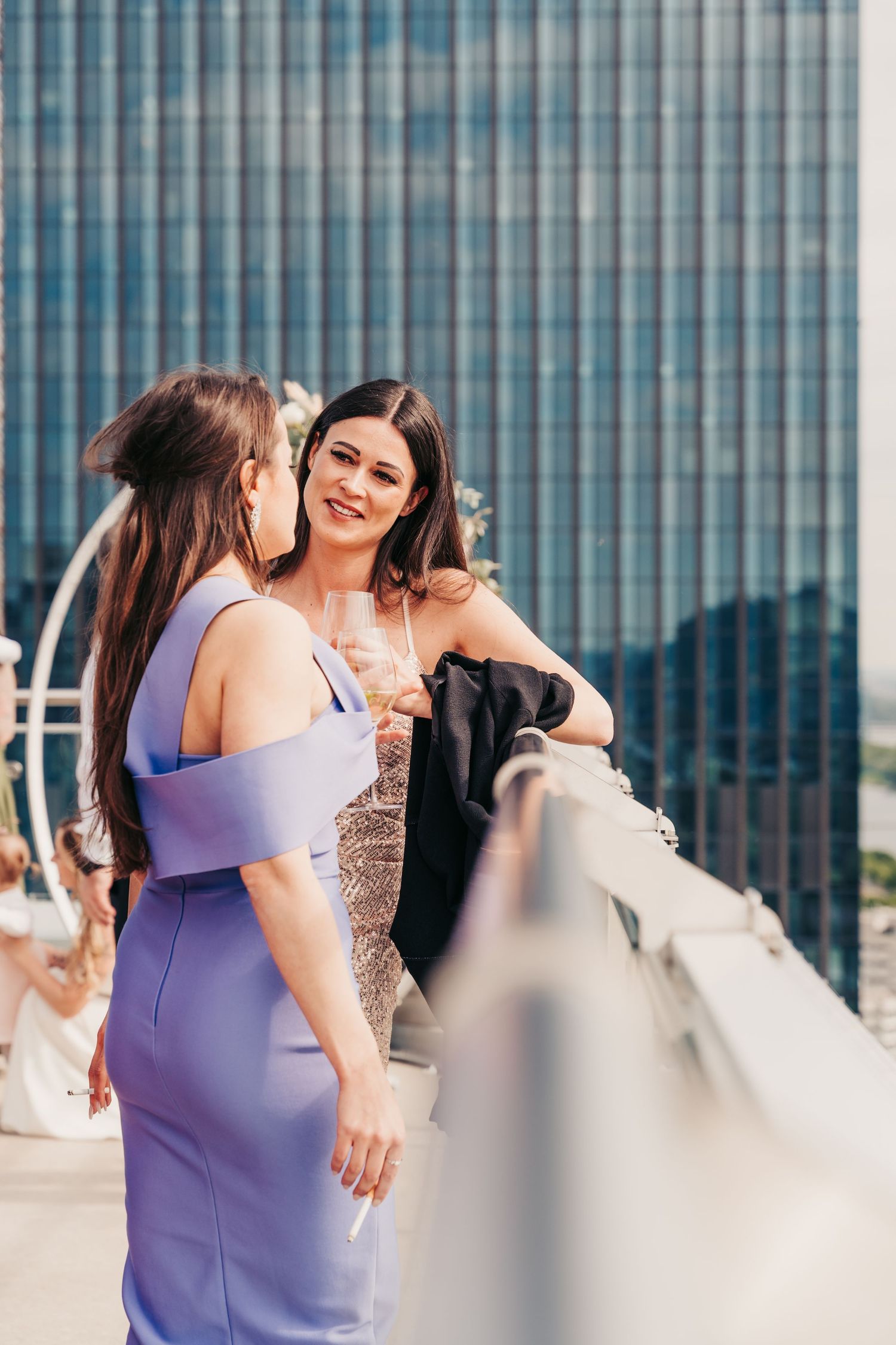 Zwei elegante Frauen in Abendkleidern halten Champagnergläser auf einer Dachterrasse mit Skyline der Stadt Wien.