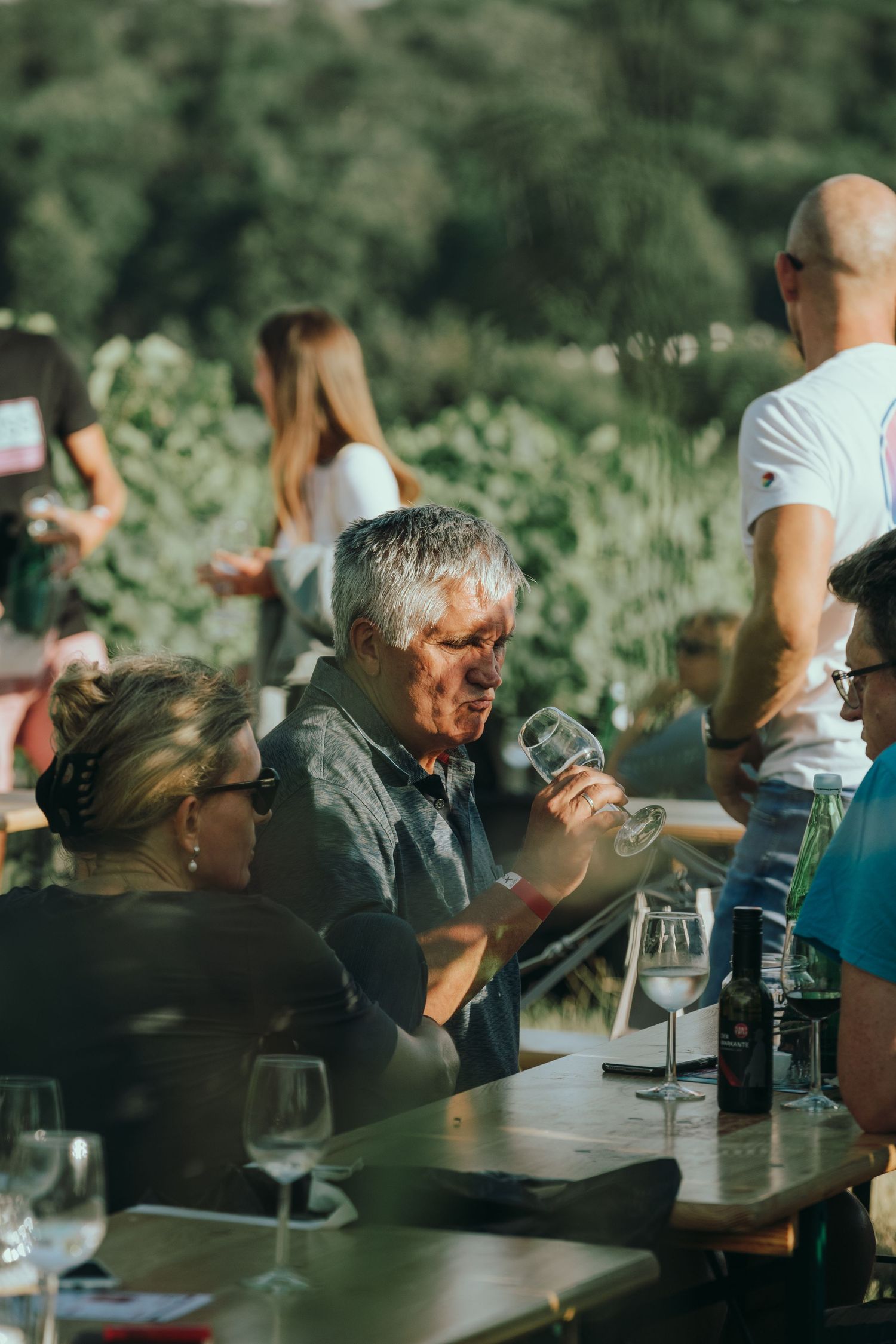 Ein älterer Mann begutachtet ein Weinglas bei einem Treffen im Freien in einem Weinberg, während Gäste an Tischen sitzen.
