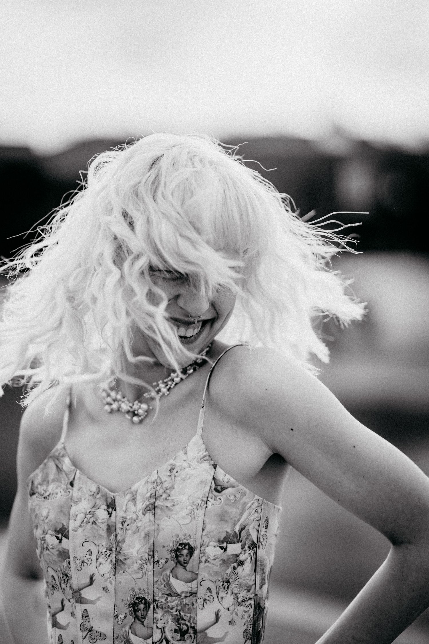 Black and white photo of a laughing woman with windswept curly blonde hair wearing a floral dress and pearl necklace.